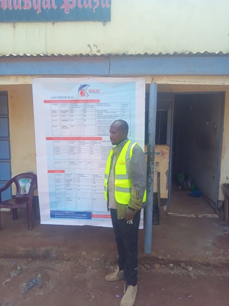 A Hikri Companion field agent beside the Last Expense Plan poster at a community registration point